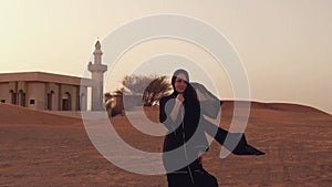 Portrait of a young Arab woman wearing traditional black clothing during beautiful sunset over the desert. Mosque on the