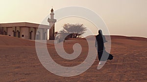 Portrait of a young Arab woman wearing traditional black clothing during beautiful sunset over the desert. Mosque on the