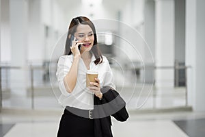 Portrait of a young Asian business woman talking over cellphone and holding cup of coffee in business building. Fashion business