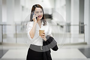 Portrait of a young Asian business woman talking over cellphone and holding cup of coffee in business building. Fashion business