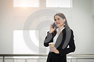 Portrait of a young Asian business woman talking over cellphone and holding cup of coffee in business building. Fashion business