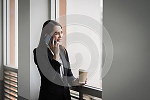 Portrait of a young Asian business woman talking over cellphone and holding cup of coffee in business building. Fashion business