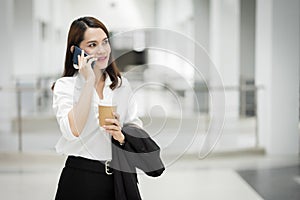 Portrait of a young Asian business woman talking over cellphone and holding cup of coffee in business building. Photo of beautiful