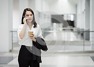 Portrait of a young Asian business woman talking over cellphone and holding cup of coffee in business building. Photo of beautiful