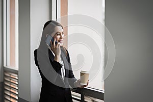 Portrait of a young Asian business woman talking over cellphone and holding cup of coffee in business building. Photo of beautiful