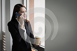 Portrait of a young Asian business woman talking over cellphone and holding cup of coffee in business building. Photo of beautiful