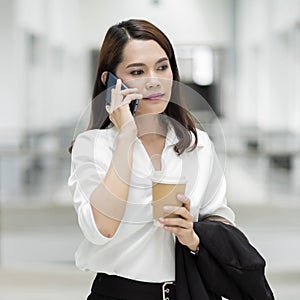 Portrait of a young Asian business woman talking over cellphone and holding cup of coffee in business building. Photo of beautiful