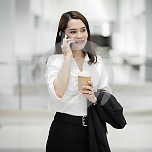 Portrait of a young Asian business woman talking over cellphone and holding cup of coffee in business building. Photo of beautiful