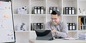 Portrait of a young Asian businessman smiling while using a laptop and writing down notes while sitting at his desk in a