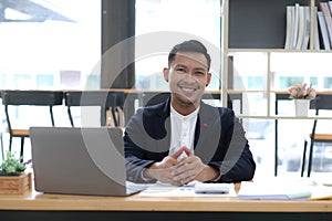 Portrait of a young Asian businessman smiling while using a laptop and writing down notes while sitting at his desk in a