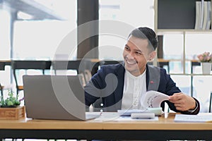 Portrait of a young Asian businessman smiling while using a laptop and writing down notes while sitting at his desk in a
