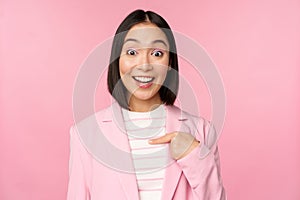 Portrait of young asian businesswoman with surprised, excited face expression, pointing finger at herself, standing in