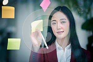 Portrait of Young asian businesswoman writing sticky paper pad on glass in modern office. Business plan concept