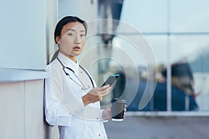Portrait young Asian doctor woman or paramedic stands exhausted on a break near the wall of a hospital clinic with a using mobile