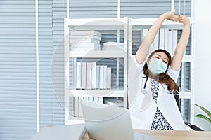 Portrait of young Asian female doctor with white coat is stretching her body while using computer laptop to check, diagnosis and