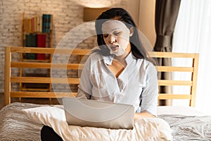 Portrait of young Asian girl applying facial mask on her face while using computer laptop in bedroom. Woman`s Beauty and