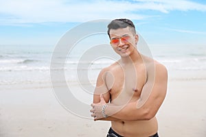 Portrait of young asian handsome and sexy man in black swimming suit and sun glasses with smiling face standing on sandy beach