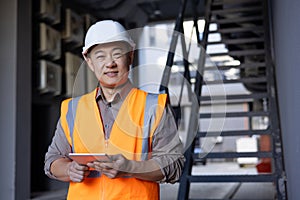 Portrait of a young Asian male builder, engineer and architect standing in a hard hat and vest outside a building