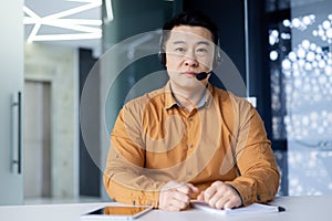 Portrait of a young Asian male businessman sitting at a desk in an office wearing a headset and looking seriously at the