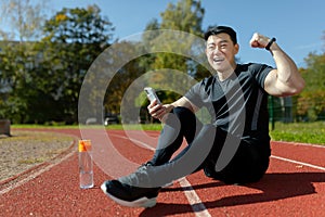 Portrait of a young Asian man, an athlete sitting on a treadmill with a phone. He is happy, looks into the camera. Shows