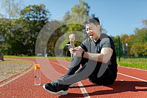 Portrait of a young Asian man, an athlete sitting on a treadmill with a phone. Rejoicing, showing a victory gesture, yes