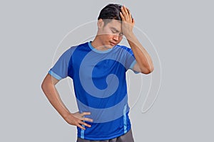 Portrait of a young Asian man with a blue t-shirt isolated on gray background, suffering from severe headache, pressing fingers to