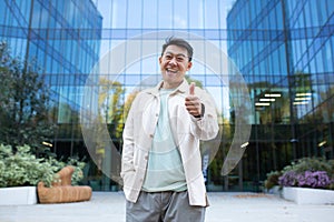 Portrait of a young Asian man standing outside an office building in casual clothes, smiling and pointing super finger