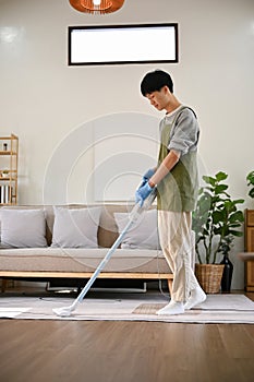 Portrait of young Asian man using stick vacuum cleaner to clean the carpet in living room