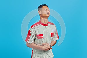 Portrait of young Asian mechanic in pain with hands on stomach over blue background