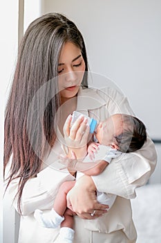 Portrait young Asian mother give milk to her newborn baby via bottle and hold in her chest in front of morning light