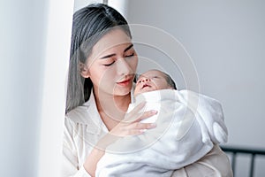 Portrait of young asian mother take care her newborn baby and holding on her chest in front of white curtain with morning light