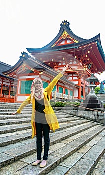 Portrait of young Asian muslim woman wearing hijab and eyeglasses standing in Fushimi Inari Shrine smiling and waving hands