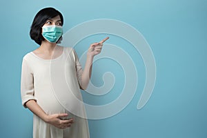 Portrait of young Asian Pregnant woman wearing medical face mask pointing to copy space on blue isolated background