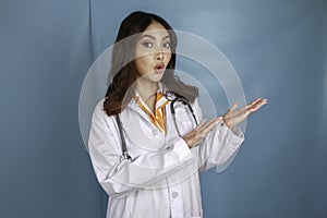 Portrait of a young Asian woman doctor, medical professional is shock and pointing at a copy space isolated over blue background