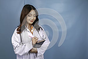 Portrait of a young Asian woman doctor, a medical professional is smiling and holding notes isolated over blue background
