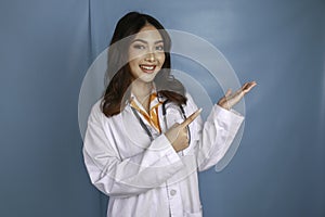 Portrait of a young Asian woman doctor, medical professional is smiling and pointing at a copy space isolated over blue background