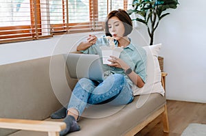 Portrait of young Asian woman freelancer eating chinese food from delivery while working on laptop in living room at home. For