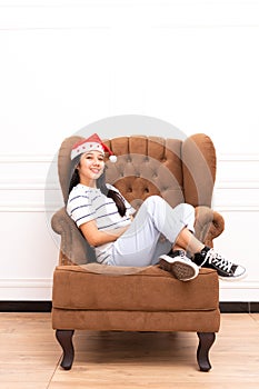 Portrait of young Asian woman wearing a Santa hat and wearing casual clothes sitting on a brown sofa