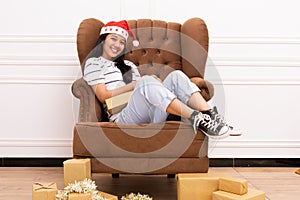 Portrait of a young Asian woman wearing a Santa hat and wearing casual clothes sitting on a brown sofa while holding a gift with a