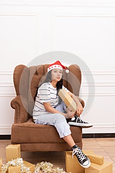 Portrait of a young Asian woman wearing a Santa hat and wearing casual clothes sitting on a brown sofa while holding a gift with a