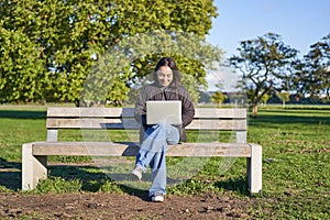 Portrait of young asian woman working on laptop, sitting on bench in park on sunny day, using her computer, studying in