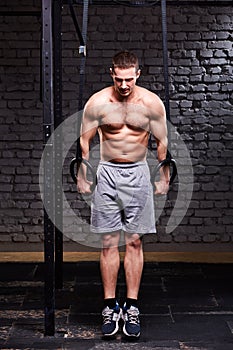 Portrait of young athlete man doing pull-ups with rings against brick wall in the cross fit gym.