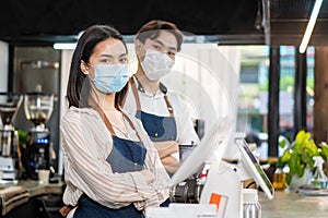 Portrait of young attractive Asian waiter and waitress wearing protective mask due to Covid-19 pandemic at cafe. Barista business