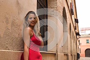 Portrait of young, attractive, blonde woman wearing an elegant red party dress and leaning against the wall of a city alley,