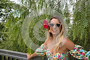 Portrait of young, attractive, blonde woman, wearing flower shirt and sunglasses, with a flower in her ear on a background of