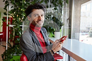 Portrait of young attractive brown haired bearded man holding smartphone in raised hand and looking gladly at camera with light