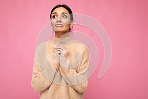Portrait of young attractive brunette woman with sincere emotions wearing beige pullover isolated over pink background