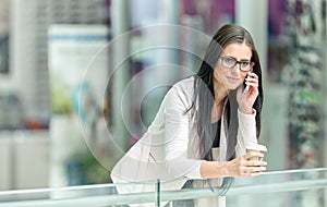 Portrait of young attractive business woman standing in the shopping mall with coffee and using her cell phone. Business break.