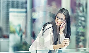 Portrait of young attractive business woman standing in the shopping mall with coffee and using her cell phone. Business break.