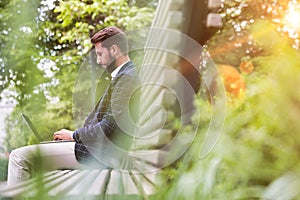 Portrait of young attractive businessman sitting on bench while working on his laptop